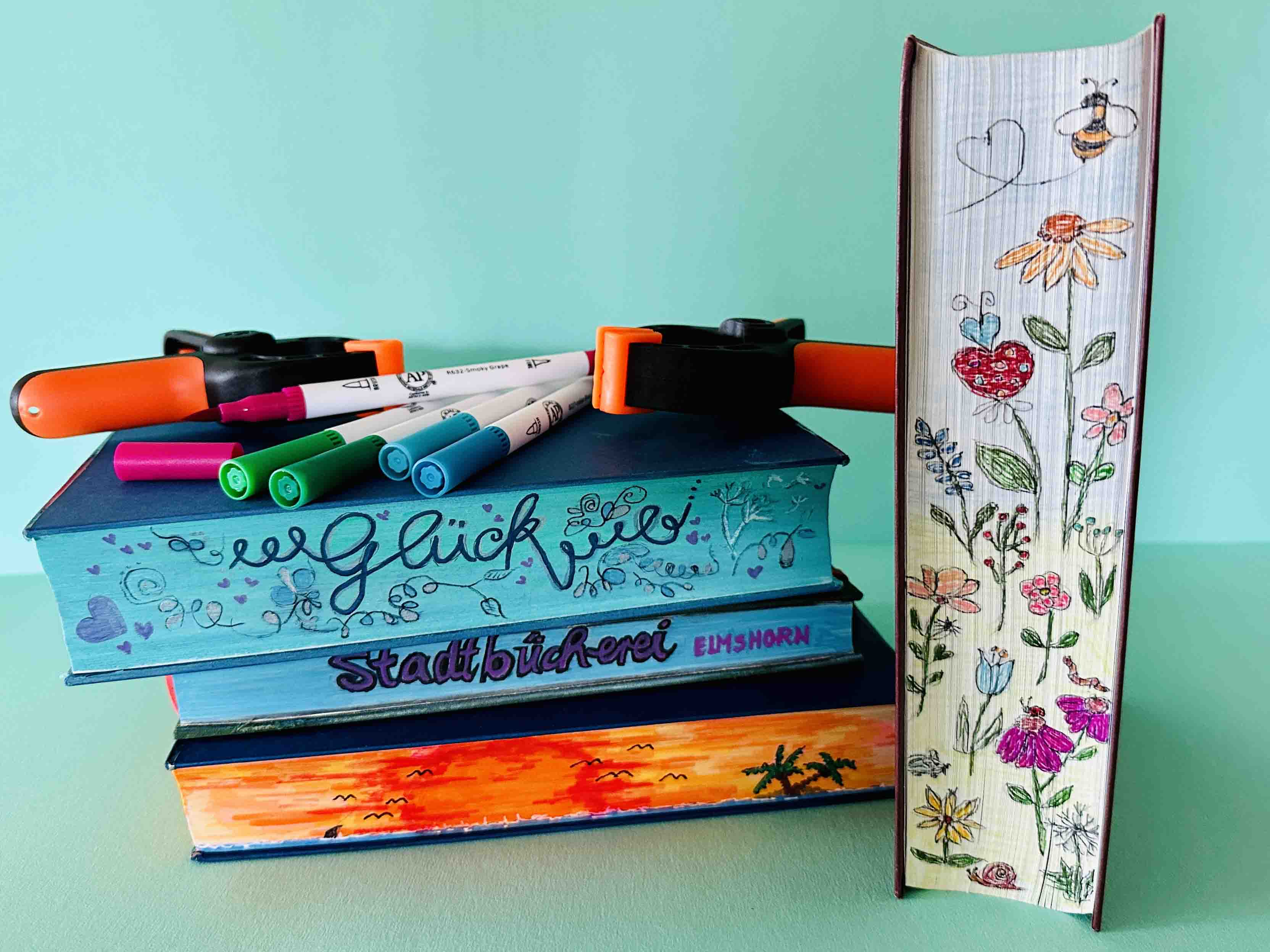 Foto Bücherstapel mit bemalten Buchkanten, auf den Büchern liegen Stifte und Buchklemmen